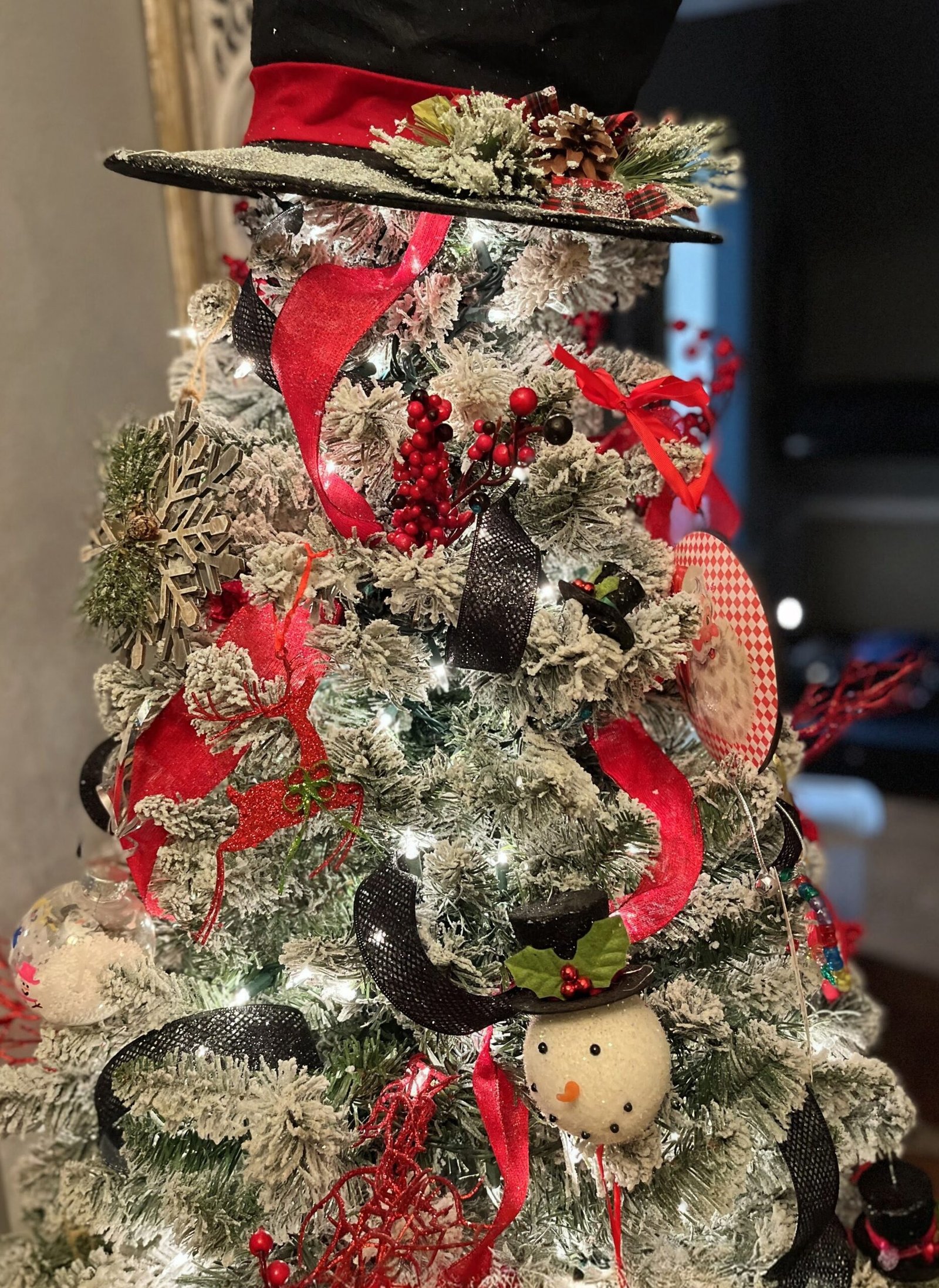 snowman christmas tree with black top hat topper, frosted branches, buffalo plaid ribbon, red berries, snowman ornaments, and warm white lights in a cozy living room